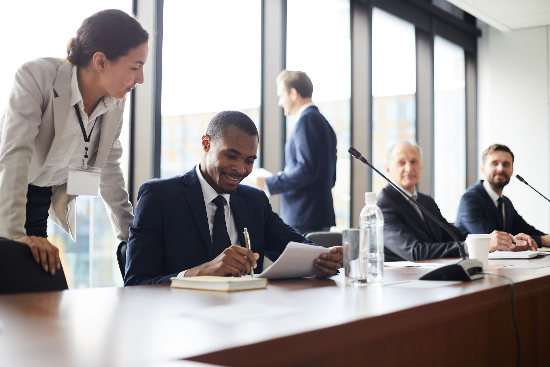 Content attractive young business lady with badge standing at black colleagues and giving advices him with papers while he analyzing information during conference break